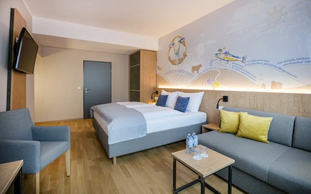 Sie sehen ein Doppelbett und eine Couch im Doppelzimmer im JUFA Hotel Weiz. Der Ort für kinderfreundlichen und erlebnisreichen Urlaub für die ganze Familie.