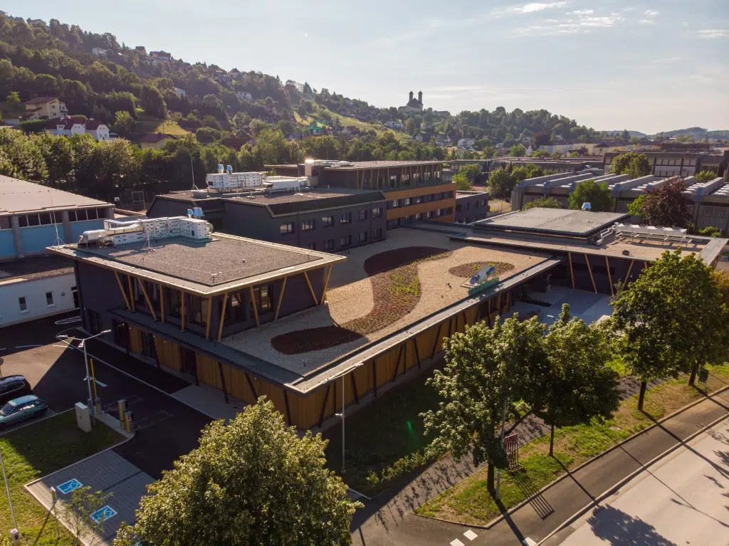 Sie sehen das JUFA Hotel Weiz in einer Luftbildaufnahme eingebettet in die Umgebung von Hügel und Bäumen.