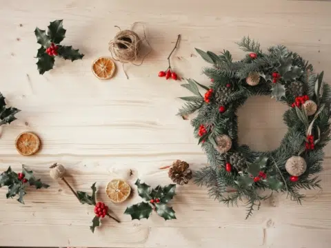 Ihr seht einen fertigen Adventkranz neben Orangenscheiben, Bindfaden und Stechpalmenzweigen.