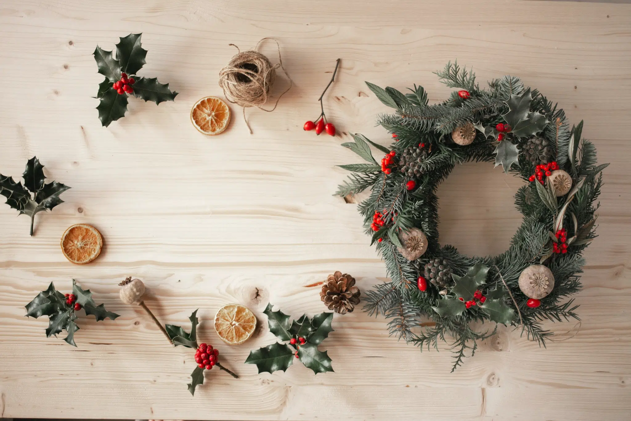 Ihr seht einen fertigen Adventkranz neben Orangenscheiben, Bindfaden und Stechpalmenzweigen.