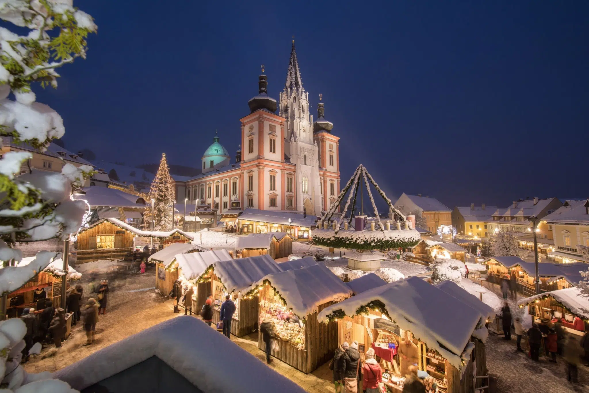 Ihr seht den verschneiten Adventmarkt in Mariazell mit der Basilika im Hintergrund und Adventmarkt-Ständen.