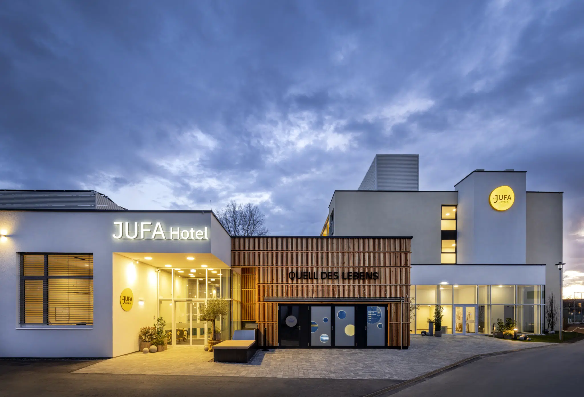 Ihr seht eine Außenansicht in der Abenddämmerung vom Haupt- und Nachteingang in das JUFA Hotel Bad Radkersburg sowie den interaktiven Erlebnisraum "Quell des Lebens" von außen.