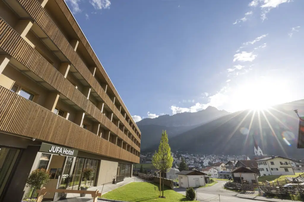 Sie sehen die wunderschöne Außenansicht des JUFA Hotel Savognin***s inklusive Bergpanorama, Dorf und Sonnenstrahlen.