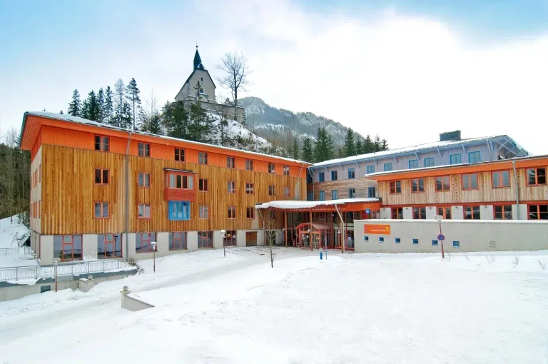 Ihr seht das JUFA Hotel Mariazell*** im Winter von außen und im Hintergrund den Sigmundsberg. Der Ort für erholsamen Familienurlaub und einen unvergesslichen Winterurlaub.