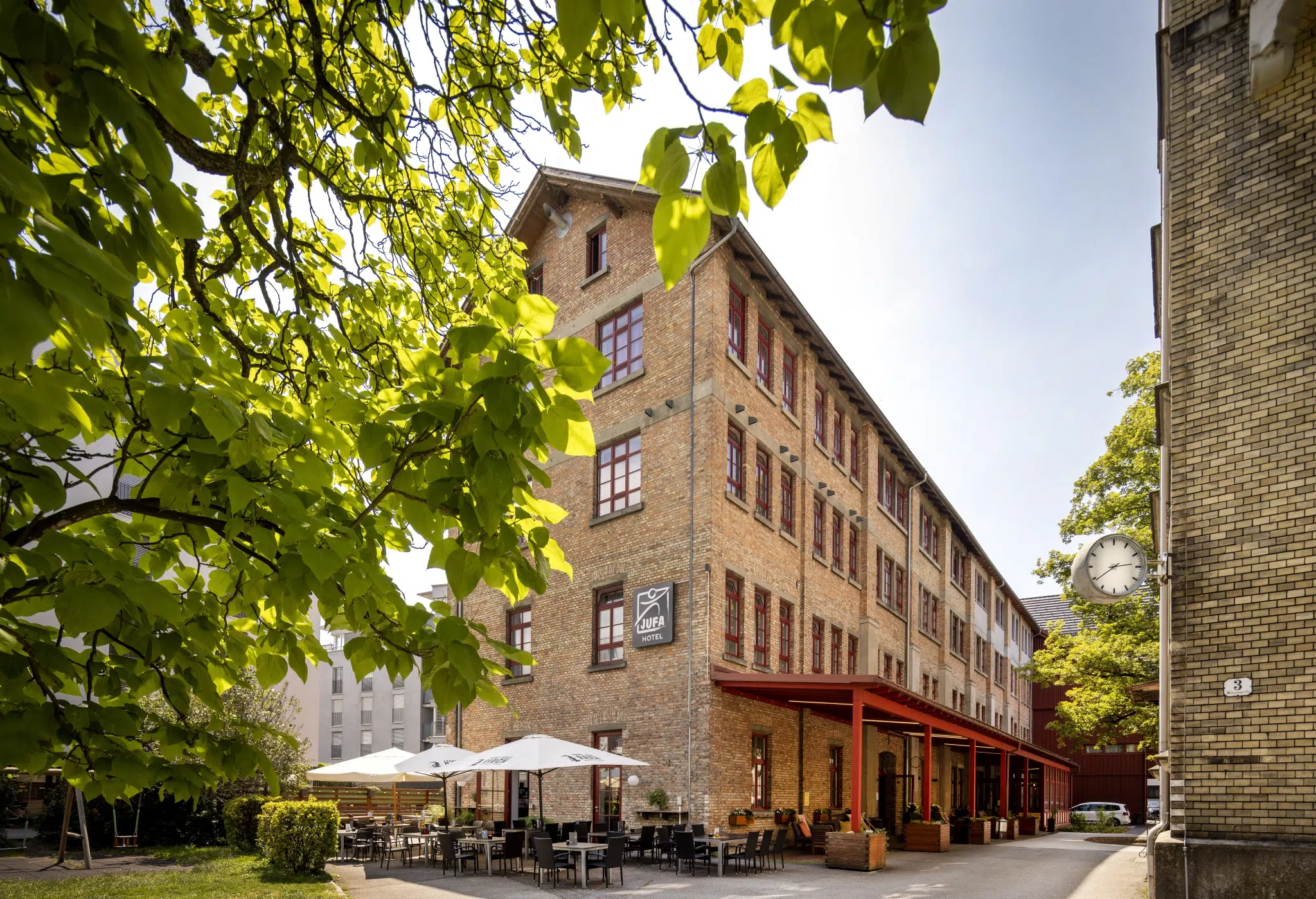 Ihr seht das historische Hauptgebäude des JUFA Hotel Bregenz am Bodensee mit Sitzgarnituren und Sonnenschirmen auf der Terrasse. Der Ort für einen unvergesslichen Wander-, Rad- und Kultururlaub für Familien, Freunde und Paare.