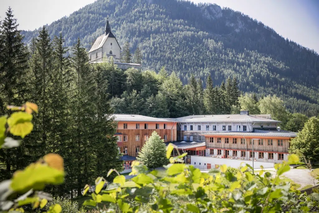 Ihr seht das JUFA Hotel Mariazell*** im Sommer von außen und im Hintergrund eine Kirche am Sigmundsberg. Der Ort für erholsamen Familienurlaub und einen unvergesslichen Winter- und Wanderurlaub.