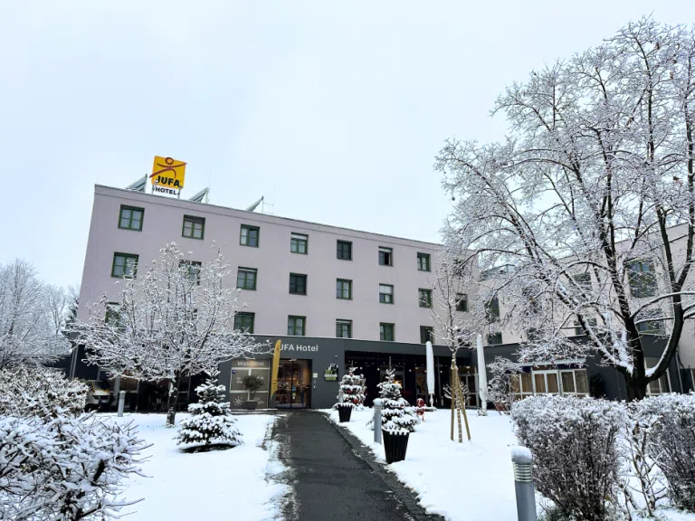 Ihr seht eine Außenansicht des JUFA Hotel Graz City*** im Winter mit Schnee und Christbäumen.