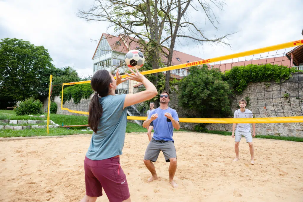 Ihr seht vier Personen beim Beachvolleyball spielen vor dem JUFA Hotel Nördlingen***. Der Ort für kinderfreundlichen und erlebnisreichen Urlaub für die ganze Familie.