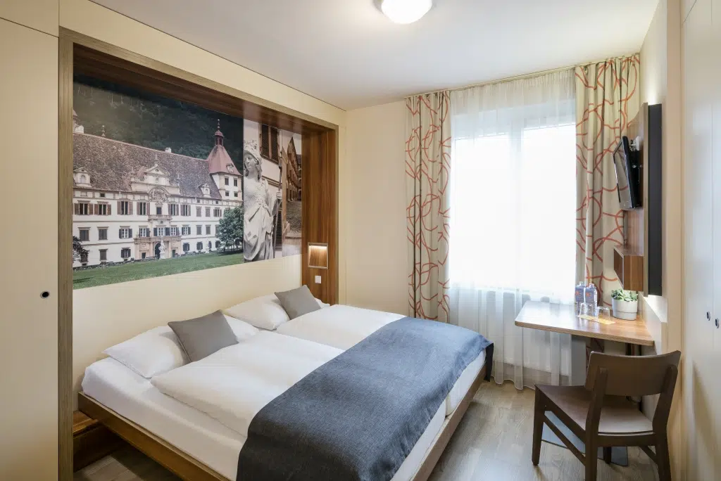 Ihr seht ein Doppelzimmer mit Bett, Kissen und Decke im JUFA Hotel Graz City***. Der Ort für erlebnisreichen Städtetrip für die ganze Familie und der ideale Platz für euer Seminar.