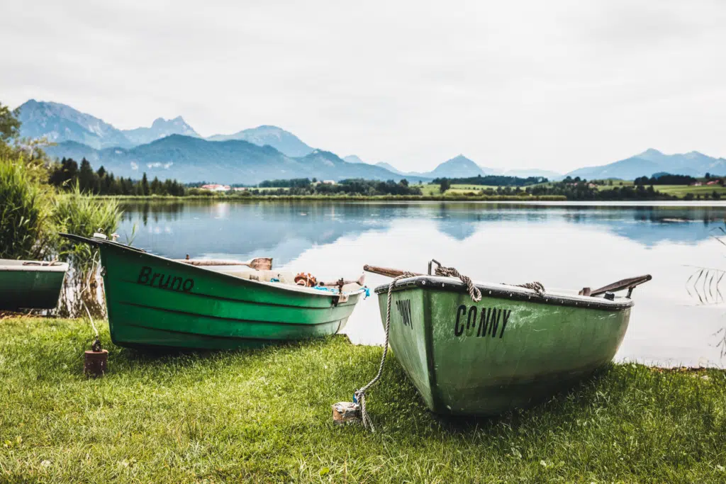 Ihr seht Boote am Ufer am Hopfensee mit Bergen im Hintergrund.