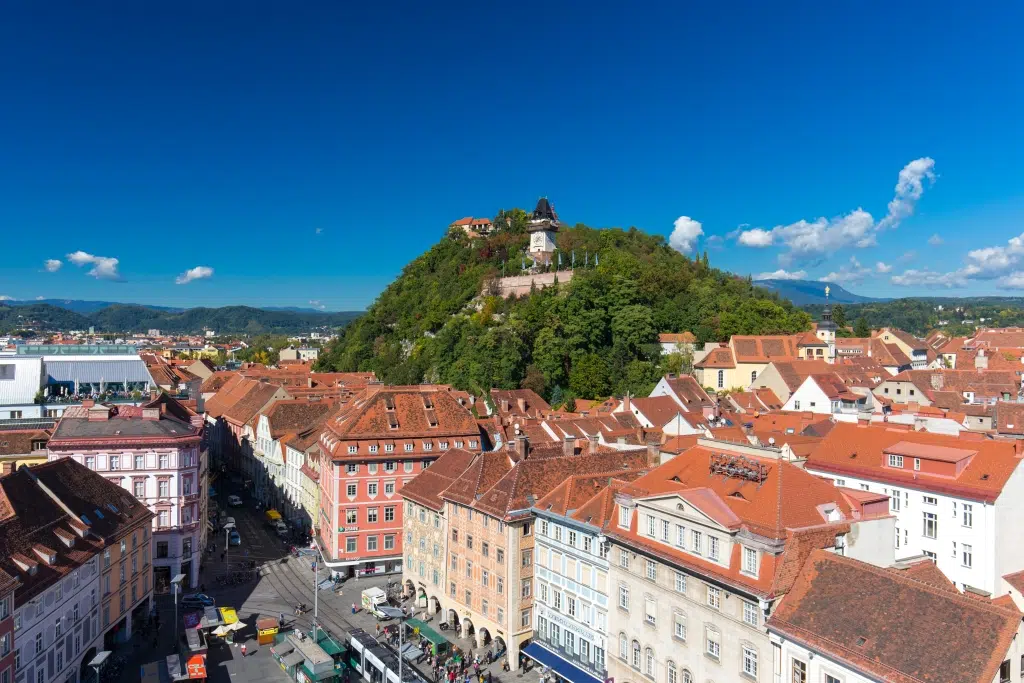 Ihr seht den Blick über die Dachlandschaft von Graz und über den Hauptplatz hin zum Schloßberg mit dem Uhrturm.