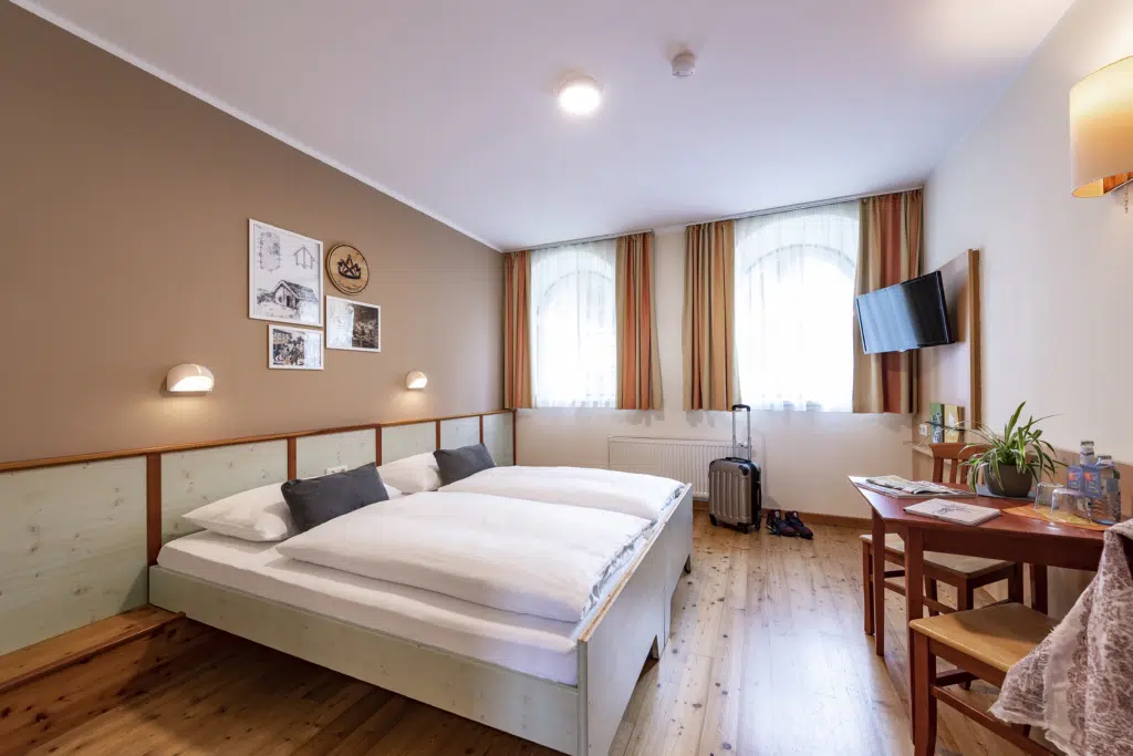 Ihr seht ein Doppelbett in einem Doppelzimmer im JUFA Hotel Murau mit TV und Tisch. JUFA Hotels bietet erholsamen Familienurlaub und einen unvergesslichen Winter- und Wanderurlaub.