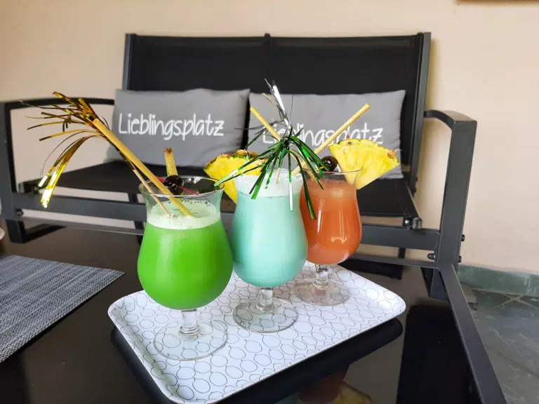 Ihr seht drei bunte köstliche Cocktails auf einem Serviertablett am Tisch, im Hintergrund liegen auf einer Sitzbank zwei Kissen mit der Aufschrift "Lieblingsplatz". JUFA Hotels bietet erholsamen Familienurlaub und einen unvergesslichen Winter- und Wanderurlaub.