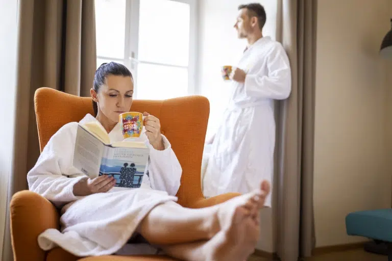 Ihr seht ein Pärchen, dass sich mit Tee und Buch am Zimmer entspannt. JUFA Hotels bietet erholsamen Familienurlaub und einen unvergesslichen Winter- und Wanderurlaub.