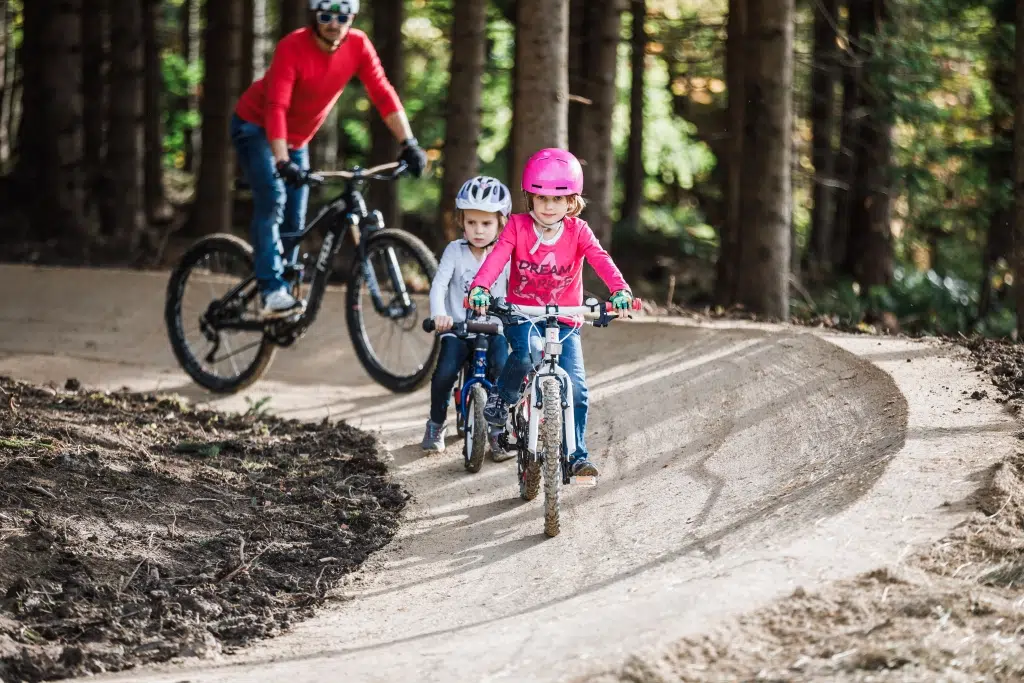 Ihr seht einen Vater mit zwei Kindern beim Mountainbiken im Mini-Bikepark beim JUFA Hotel Annaberg***s. Der Ort für erholsamen Familienurlaub und einen unvergesslichen Winter- und Wanderurlaub.