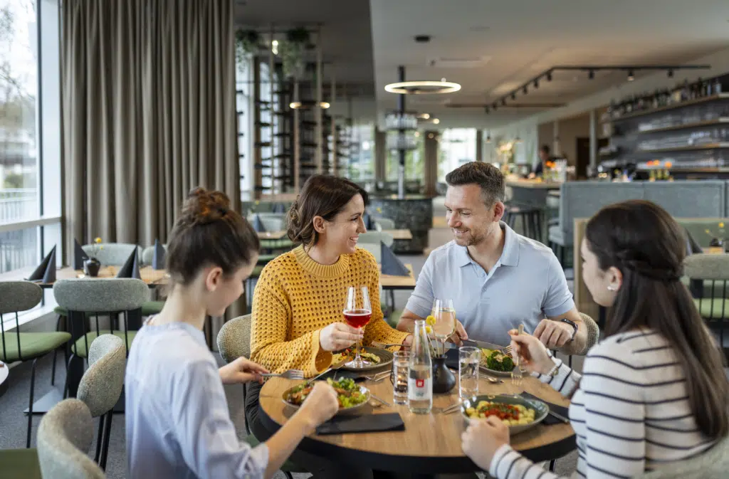 Sie sehen eine vierköpfige Familie beim gemütlichen Mittag- und Abendessen mit regionalen Speisen und Getränken im Café & Restaurant Wandelhalle im JUFA Hotel Bad Radkersburg