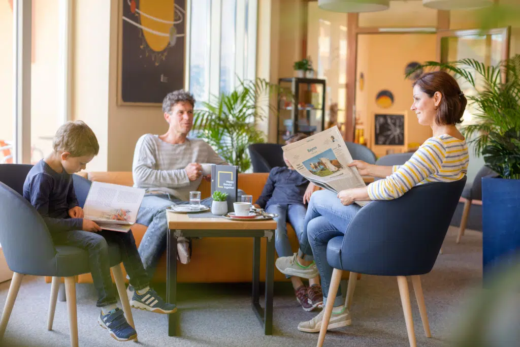 Ihr seht eine Familie mit zwei Kindern bei der Nachmittagspause mit Buch und Zeitung im JUFA Hotel Nördlingen***. Der Ort für kinderfreundlichen und erlebnisreichen Urlaub für die ganze Familie.