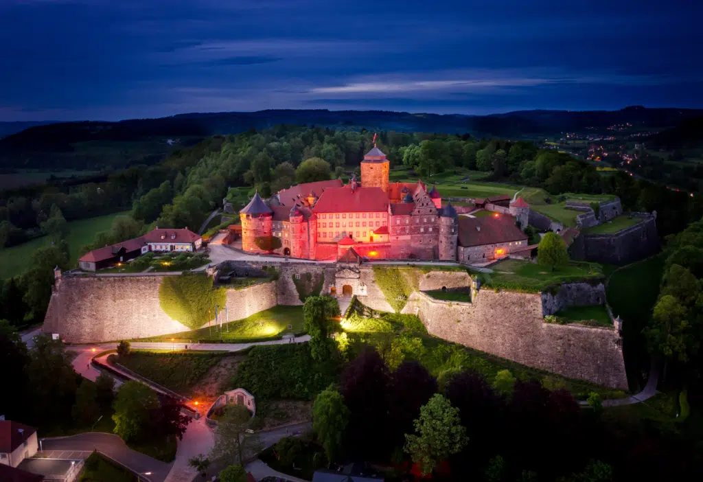 Ihr seht die Festung Rosenberg in Kronach mit Lichtern am Abend. JUFA Hotels bietet kinderfreundlichen und erlebnisreichen Urlaub für die ganze Familie.