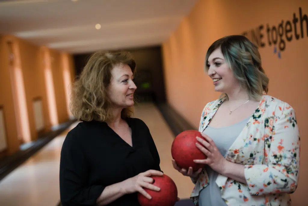 Ihr seht zwei Frauen mit roten Bowlingkugeln auf der Kegelbahn im JUFA Hotel Stubenbergsee. Der Ort für erholsamen Familienurlaub und einen unvergesslichen Winterurlaub.