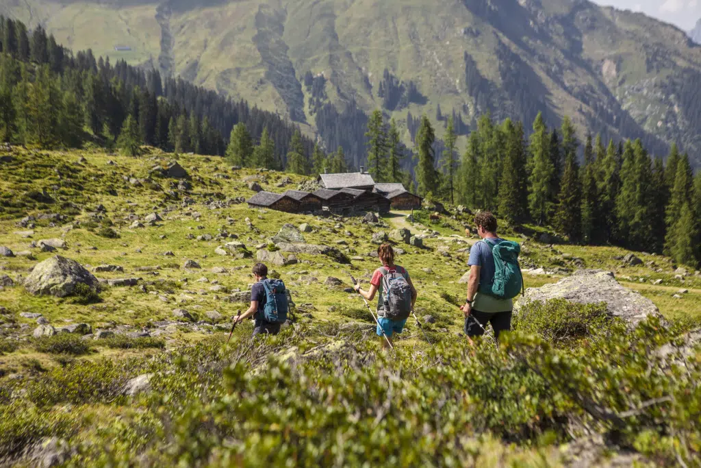 Sie sehen eine Familie die entlang einiger Almhütten zur Alpguesalpe wandert. JUFA Hotels bietet kinderfreundlichen und erlebnisreichen Urlaub für die ganze Familie.