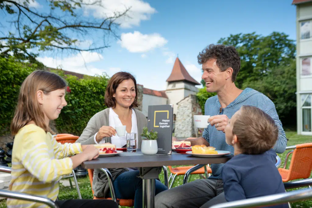 Ihr seht eine Familie beim Nachmittagskaffee mit Kuchen im Garten im JUFA Hotel Nördlingen***. Der Ort für kinderfreundlichen und erlebnisreichen Urlaub für die ganze Familie.