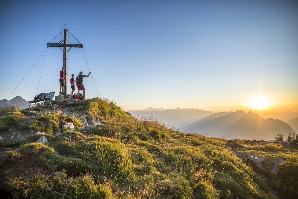 Sie sehen drei Freunde, die den Gipfel des Muttjöchles erreicht haben und den beginnenden Sonnenaufgang genießen. JUFA Hotels bietet Ihnen den Ort für erlebnisreichen Natururlaub für die ganze Familie.