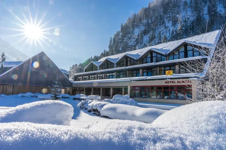 Ihr seht eine Außenansicht vom JUFA Hotel Grünau im Almtal mit Sonnenschein im Winter. JUFA Hotels bietet erholsamen Familienurlaub und einen unvergesslichen Winterurlaub.