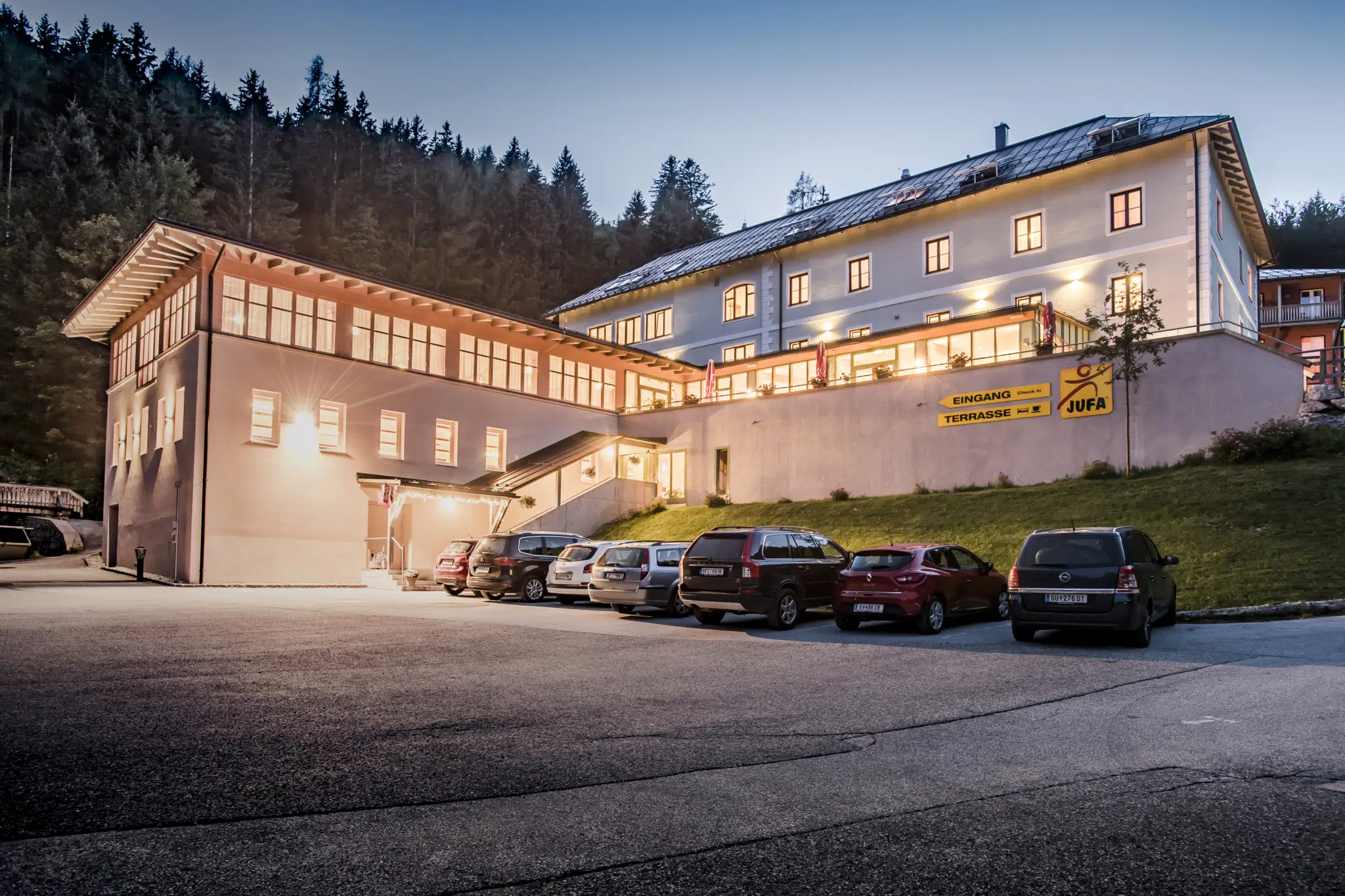 Außenansicht vom JUFA Hotel Altaussee mit Parkplatz bei Abendstimmung. JUFA Hotels bieten erholsamen Familienurlaub und einen unvergesslichen Winter- und Wanderurlaub.