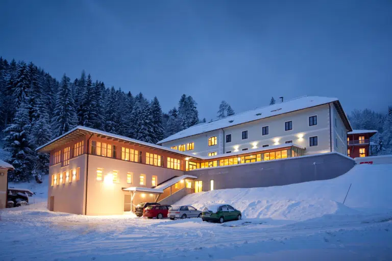 Außenansicht vom JUFA Hotel Altaussee mit Parkplatz bei Abendstimmung im Winter. JUFA Hotels bietet erholsamen Familienurlaub und einen unvergesslichen Winterurlaub.