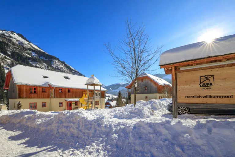 Ihr seht eine Außenansicht vom JUFA Hotel Donnersbachwald - Almerlebnis mit schneebedecktem Willkommensschild im Winter. JUFA Hotels bietet erholsamen Familienurlaub und einen unvergesslichen Winterurlaub.