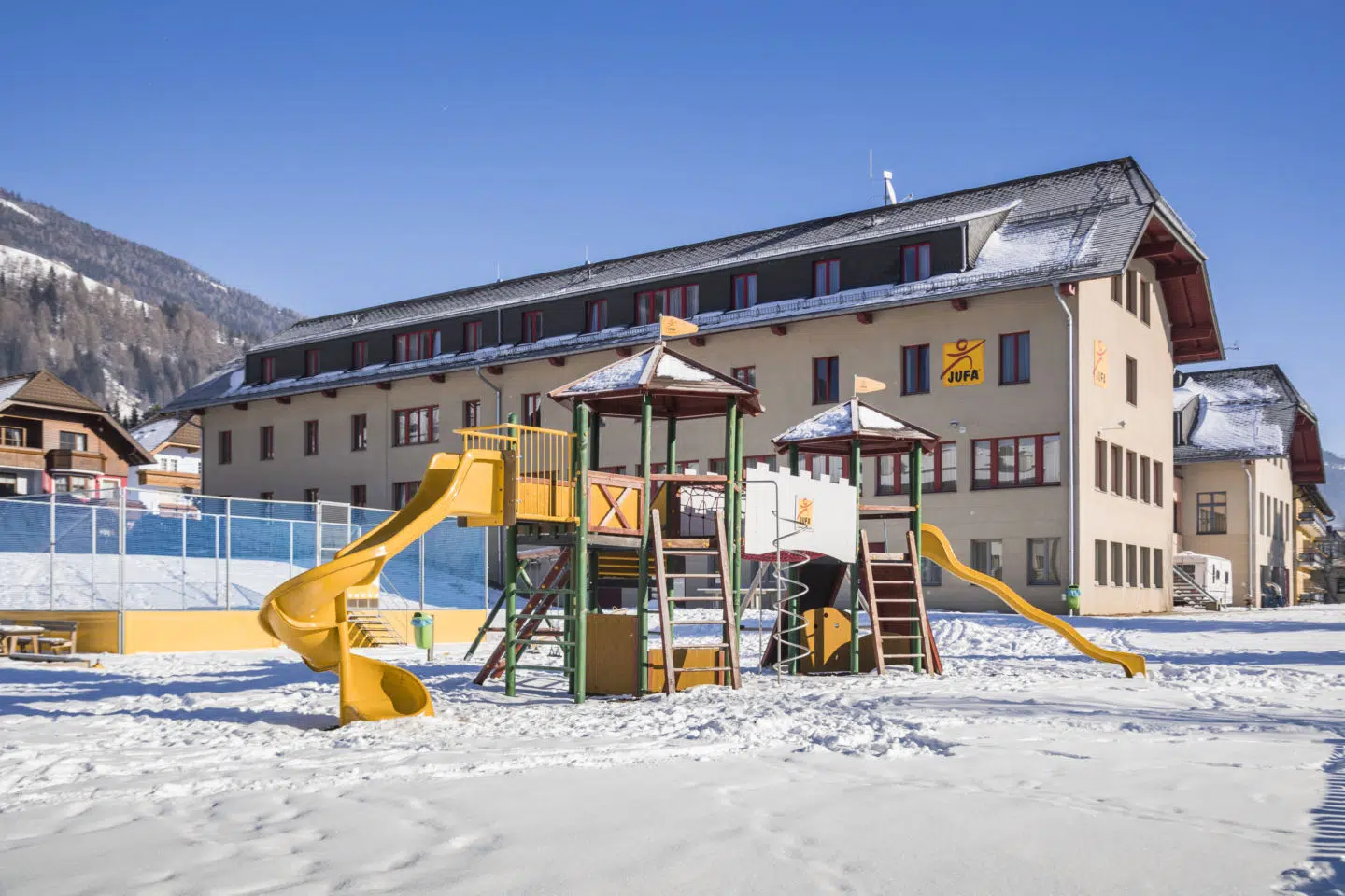 Ihr seht eine Außenansicht vom JUFA Hotel Lungau mit Spielplatz im Winter.