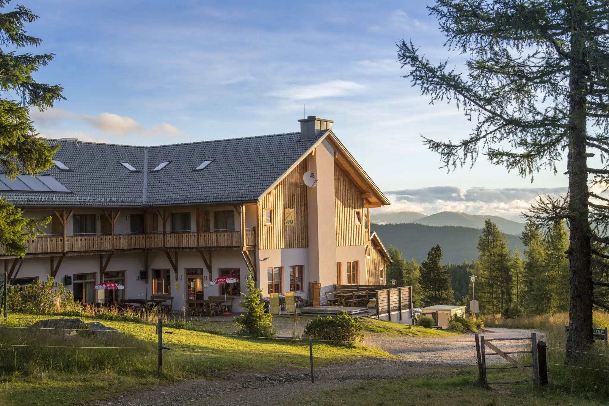 Außenansicht mit Anfang des Wanderwegs im Sommer vom JUFA Hotel Nockberge Almerlebnis. Der Ort für erholsamen Familienurlaub und einen unvergesslichen Winter- und Wanderurlaub.