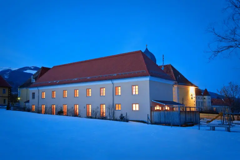 Ihr seht die Außenansicht im Winter des Hotels mit beleuchteten Fenstern bei Abendstimmung beim JUFA Hotel Seckau. Der Ort für erholsamen Familienurlaub und einen unvergesslichen Winter- und Wanderurlaub.