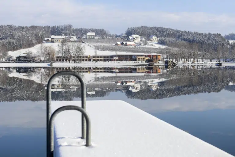 Ihr seht eine Außenansicht vom JUFA Hotel Stubenbergsee mit See und verschneitem Steg im Winter. JUFA Hotels bietet kinderfreundlichen und erlebnisreichen Urlaub für die ganze Familie.