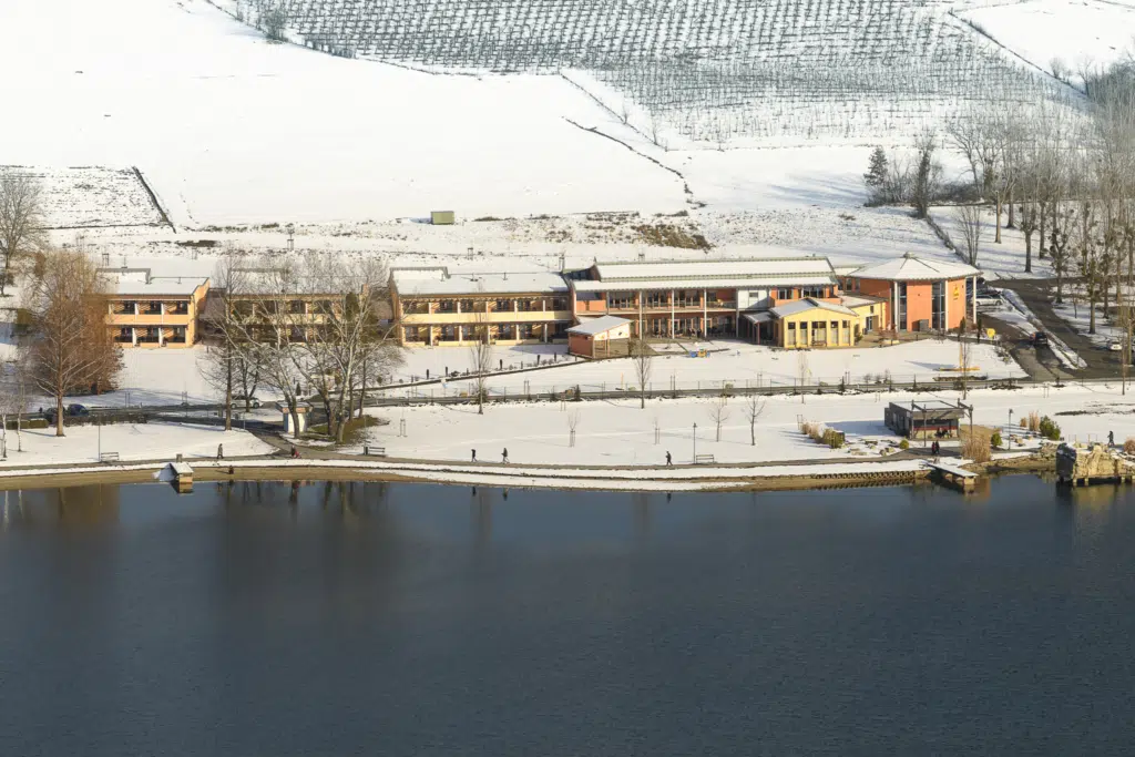 Ihr seht eine Außenansicht vom JUFA Hotel Stubenbergsee*** mit Seeblick im Winter. JUFA Hotels bietet erholsamen Familienurlaub und einen unvergesslichen Winterurlaub.