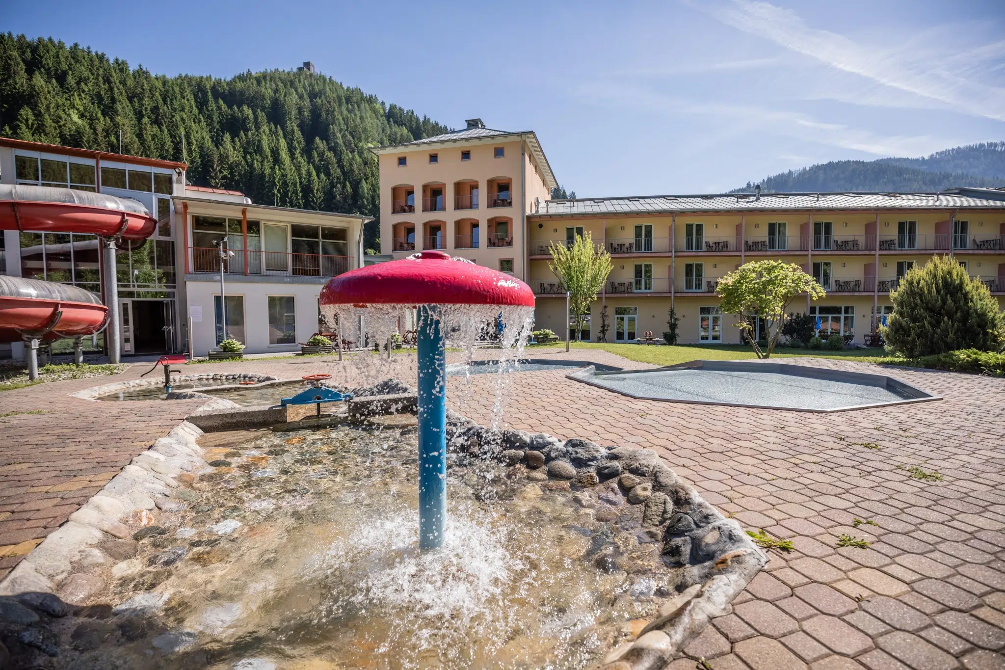 Sie sehen eine Außenansicht vom JUFA Hotel Veitsch mit Pilzbrunnen und Kinderbecken im Sommer. JUFA Hotels bietet kinderfreundlichen und erlebnisreichen Urlaub für die ganze Familie.