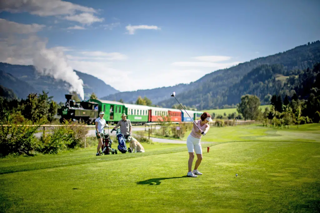 Ihr seht drei Personen mit einem Hund beim Golfen in der Region Murau. Im Hintergrund seht ihr einen vorbeifahrenden Zug der Murtalbahn, welche euch komfortabel ins JUFA Hotel Murau*** bringt.