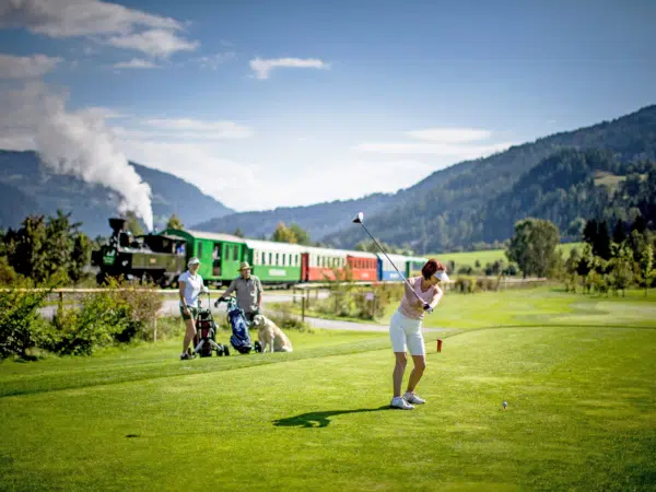 Ihr seht drei Personen mit einem Hund beim Golfen in der Region Murau. Im Hintergrund seht ihr einen vorbeifahrenden Zug der Murtalbahn, welche euch komfortabel ins JUFA Hotel Murau*** bringt.