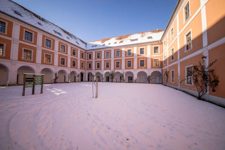 Ihr seht den schneebedeckten Innenhof vom JUFA Hotel mit Wegweiser im Winter. JUFA Hotels bietet den Ort für erfolgreiche und kreative Seminare in abwechslungsreichen Regionen.