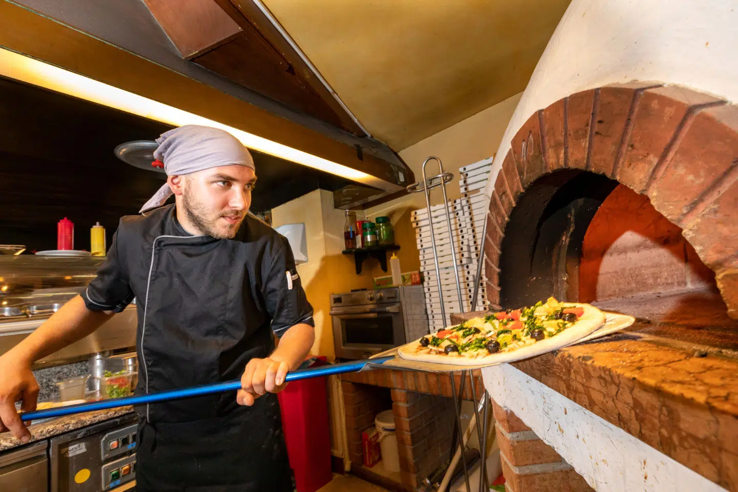 Ihr seht einen Pizzakoch, der eine Pizza in den Holzofen schiebt.