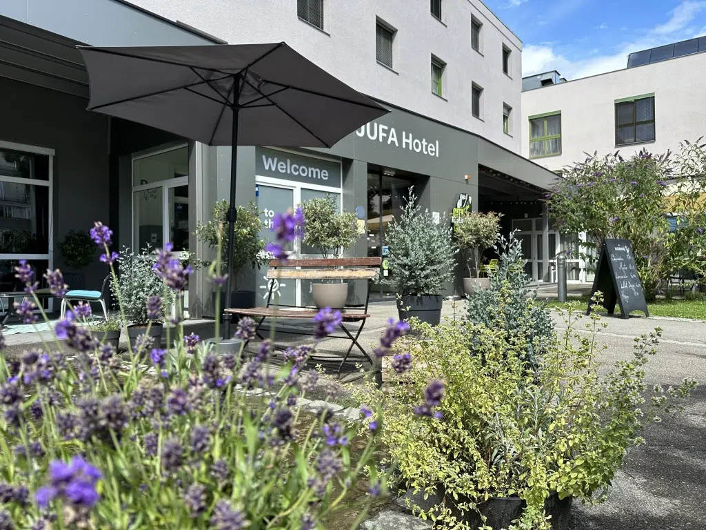 Ihr seht die Terrasse und den Eingang des JUFA Hotel Graz City mit Stühlen, Lehnsesseln, Cafétischen und Sonnenschirmen, im Vordergrund ein blühender Lavendelbusch. Der Ort für einen erlebnisreichen Städtetrip für die ganze Familie und der ideale Platz für euer Seminar.