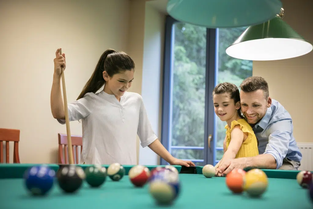 Ihr seht einen Vater mit seinen Kindern beim Billard spielen im JUFA Hotel Mariazell***. Der Ort für kinderfreundlichen und erlebnisreichen Urlaub für die ganze Familie.