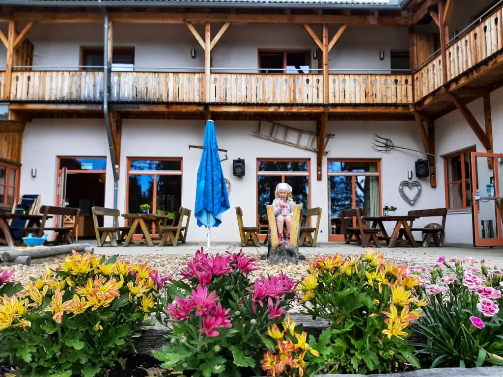 Ihr seht die Terrasse mit bunten Blumen, im Hintergrund sitzt ein Mädchen mit Lollipop. JUFA Hotels bietet erholsamen Ski- und Wanderurlaub für Familien in Kärnten.