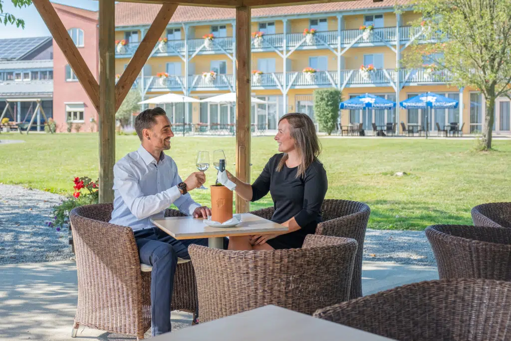 Ihr seht ein Pärchen, das mit Wein anstößt, unter dem Pavillon im Garten des JUFA Hotel Vulkanland. Der Ort für erholsamen Familienurlaub und einen unvergesslichen Winter- und Wanderurlaub.