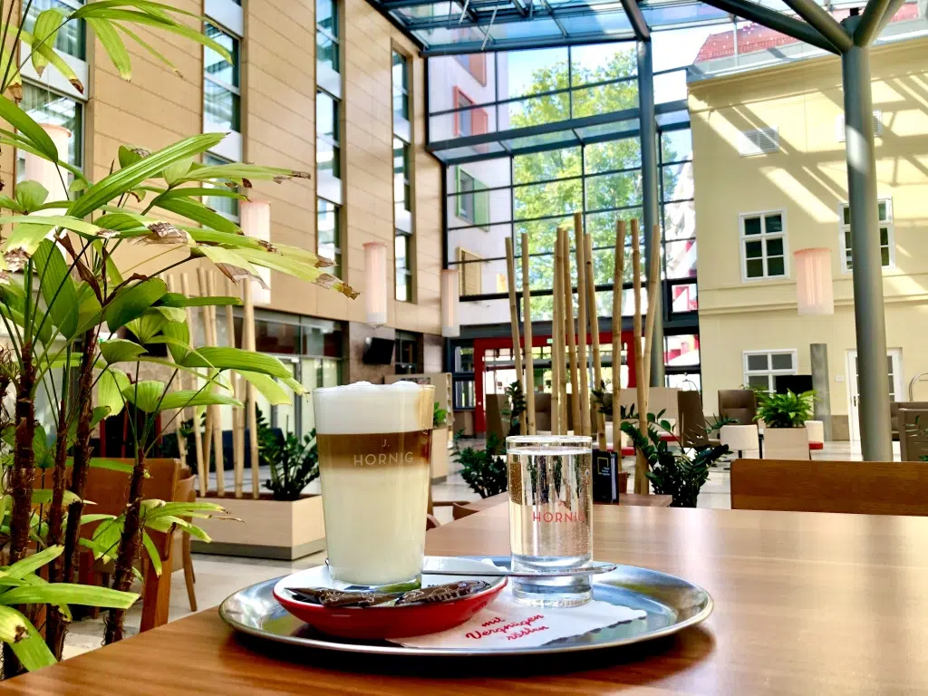 Ihr seht ein Glas mit Latte Macchiato sowie ein Wasserglas auf einem Tablett, das auf einem Tisch in der Lobby des JUFA Hotel Wien City**** steht. Der Ort für erholsamen Städteurlaub und bestens betreute Seminare in Österreich.