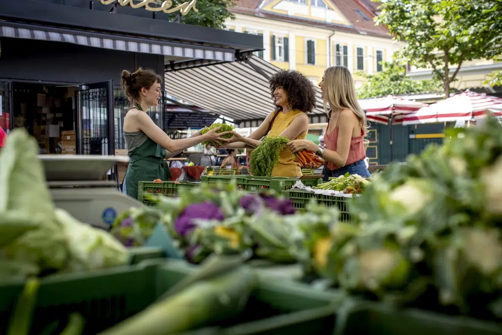 Ihr seht zwei junge Frauen, die am Kaiser-Josef-Markt in Graz bei einer Frau Gemüse kaufen.