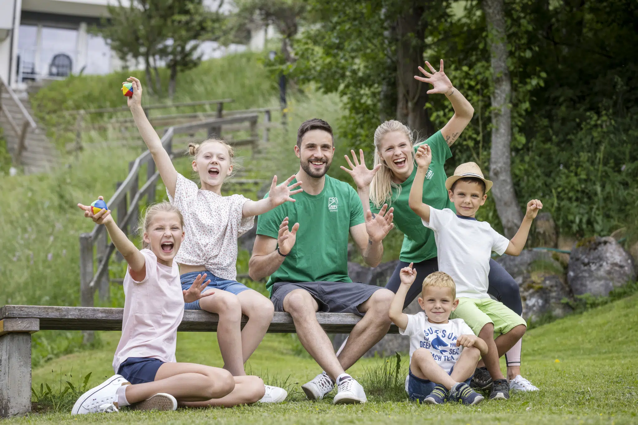 Ihr seht zwei Kids-Club Betreuer der JUFA Hotels und Kinder auf einer Bank sitzen und Spaß haben.