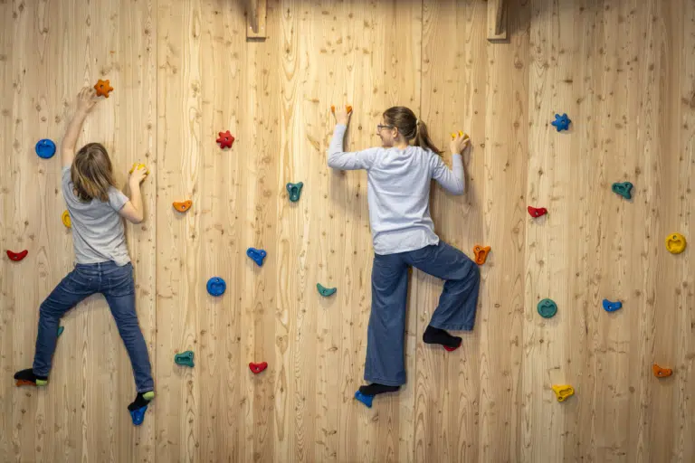 Ihr seht zwei Kinder, die gerade die hölzerne Boulderwand im Indoor-KInderspielbereich im JUFA Hotel Bad Radkersburg erklimmen.