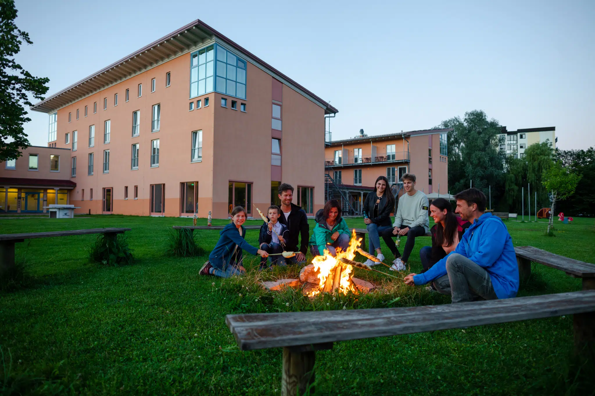 Ihr seht die Hausansicht mit Lagerfeuerstelle im Vordergrund im JUFA Familienresort Kempten***s. Der Ort für kinderfreundlichen und erlebnisreichen Urlaub für die ganze Familie.