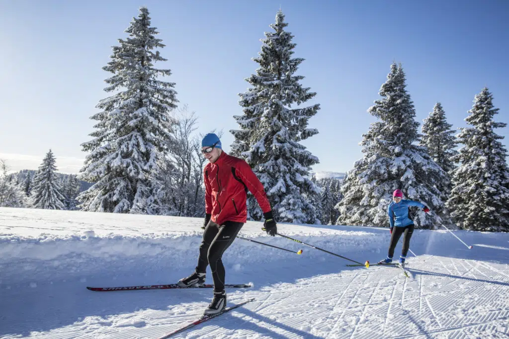 Ihr seht 2 Personen beim Langlaufen auf der Loipe Herzogenhorn mit winterlicher Landschaft im Hintergrund.